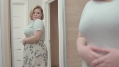 Genç obez kadın şort deniyor, aynaya bakıyor, kendine güveni yok.