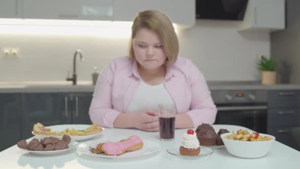 Задумчивая толстая женщина смотрит на торты и пиццу на столе, потеря веса — стоковое видео