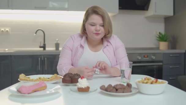 Толстушка пьет таблетки с водой, смотрит на сладкие десерты на столе — стоковое видео