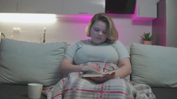 Mosolygó fiatal nő olvasás érdekes könyvet, borított takaró a kanapén — Stock videók