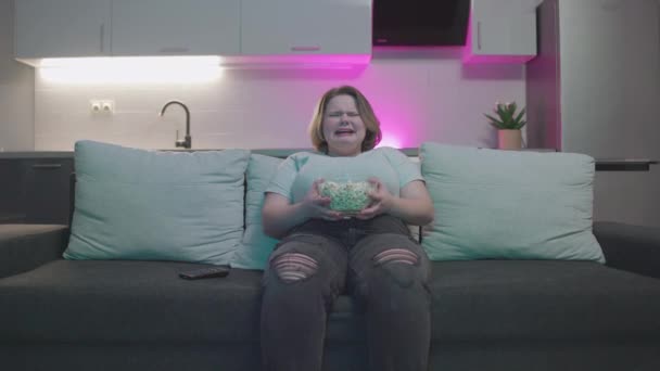 Плачущая женщина смотрит драму по телевизору и ест попкорн, сидит на диване, отдыхает — стоковое видео
