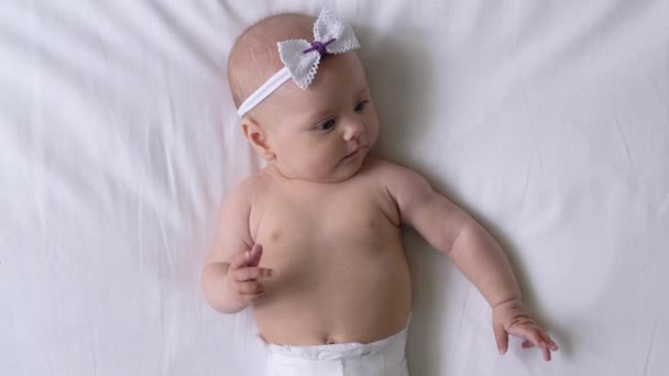 Grappig baby meisje in vlinder knoop op hoofd liggend op bed, comfort in luier — Stockvideo