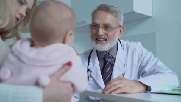 Доброзичливий старший лікар, який вивчає дитячий горло, консультує молоду маму в клініці — стокове відео