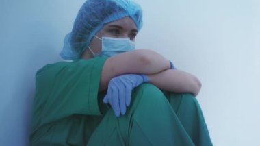 Koruyucu maskeli ve eldivenli üzgün doktor hastanede yerde oturuyor.