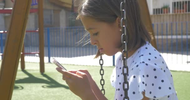 スマート フォンで遊んで 子供の遊び場の携帯電話スマート フォンの新しい技術を使用してかわいい美しい女の子 — ストック動画