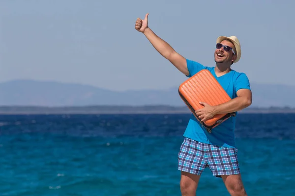 Muž drží zavazadla a ukazuje palec proti modrému oceánu. Cestovní koncept — Stock fotografie