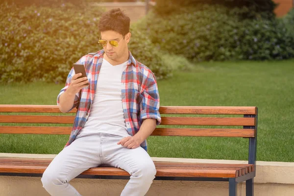 Mladý muž sedí na lavičce a pomocí smartphone v parku — Stock fotografie