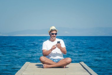 İskelede oturup ve smartphone kullanarak turist adam. Yaz ve