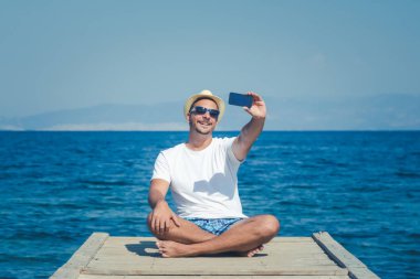 Şapka ve güneş gözlüğü iskele ve alma selfi oturan genç adam