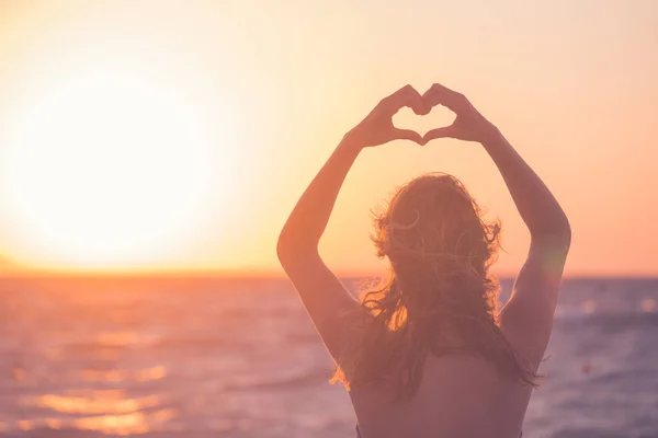 Mujer haciendo forma de corazón con sus manos al atardecer sobre el mar —  Fotos de Stock