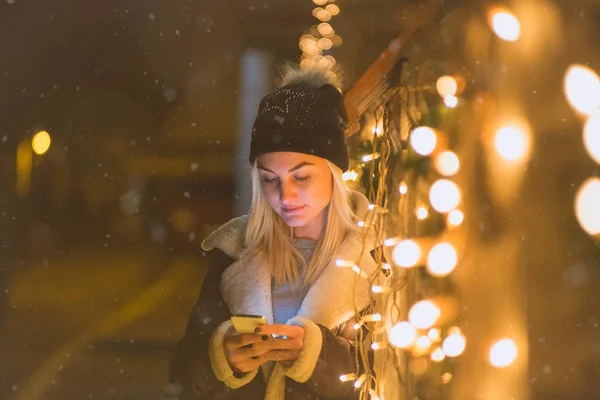 Молода жінка використовує смартфон поруч з різдвяними вогнями на вулиці — стокове фото