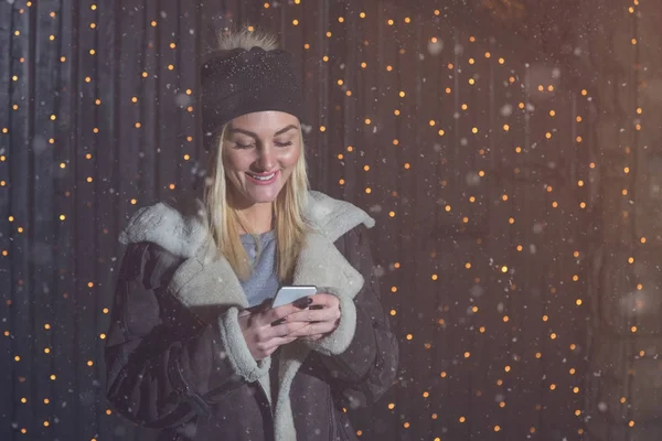 Mujer joven usando el teléfono inteligente junto a las luces de Navidad en el st — Foto de Stock