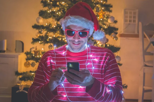 Homme avec chapeau de Père Noël et lumières de Noël en utilisant smartphone. Arbre de Noël en arrière-plan — Photo