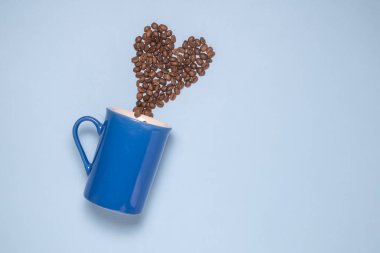 Pastel mavi zemin üzerine mavi kupa ve kahve çekirdekleri kalp şeklinde yüksek açılı görünüş.