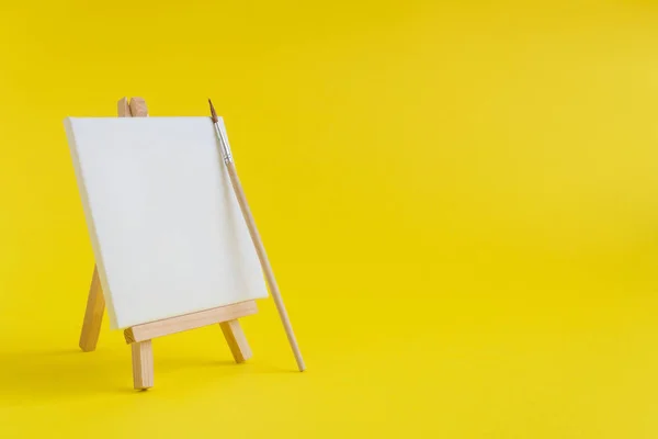 Leere Leinwand mit Holzständerminiatur und Pinsel auf schlichtem gelben Hintergrund. Kunstausstattung minimalistisches Konzept. — Stockfoto