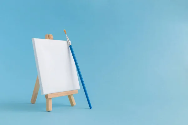 Leere Leinwand mit Holzständerminiatur und Pinsel auf schlichtem blauem Hintergrund. Kunstausstattung minimalistisches Konzept. — Stockfoto