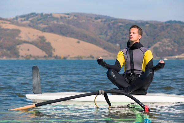 Homem de fato de mergulho sentado na prancha de surf em posição de lótus e fazendo exercícios de meditação zen. Windsurf e conceito de equilíbrio de vida . — Fotografia de Stock