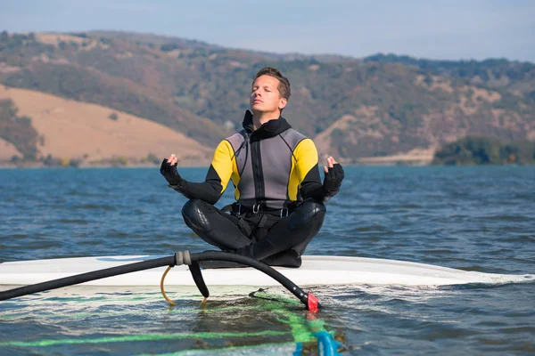 Homem de fato de mergulho sentado na prancha de surf em posição de lótus e fazendo exercícios de meditação zen. Windsurf e conceito de equilíbrio de vida . — Fotografia de Stock