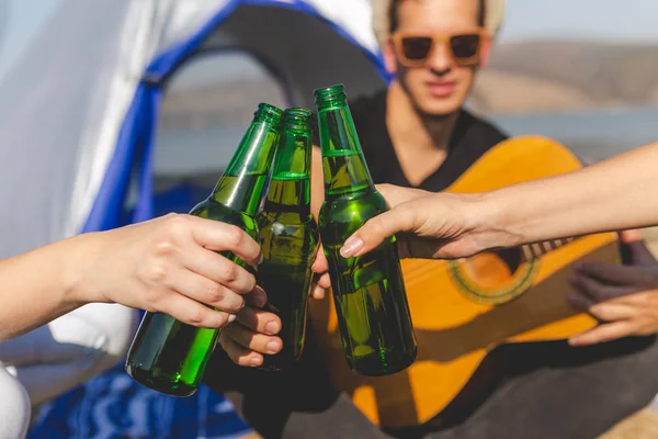 Крупный план молодого человека с гитарой перед палаткой на пляже, держащего пиво и болеющего со своими друзьями . — стоковое фото