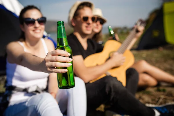 Крупный план веселой женщины, держащей бутылку пива, сидя с друзьями перед палаткой на природе. Озил и концепция свободы . — стоковое фото
