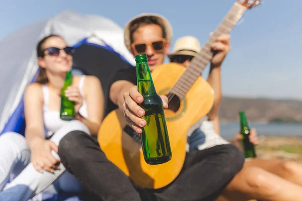 Гитарист показывает бутылку пива, развлекаясь со своими друзьями во время похода на пляже . — стоковое фото
