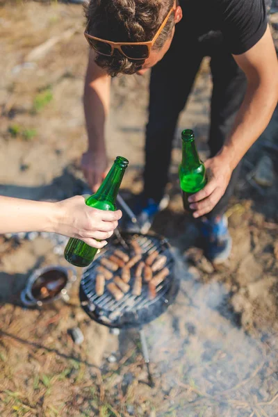 Friends clinking bottles of beer while preparing kebab lunch on barbecue grill outdoors.