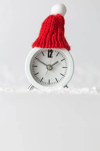 White alarm clock wearing red winter hat in snow minimal creative christmas holiday concept. — Stock Photo, Image