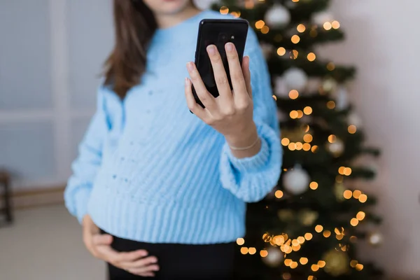 Close up of young pregnant woman holding mobile phone against christmas tree at home.