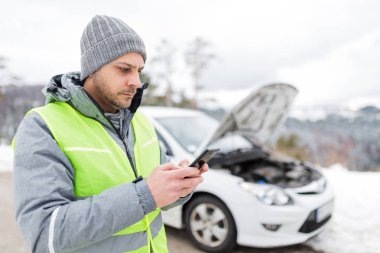 Akıllı telefon kullanan ve yol yardımı acil servisini arayan bir adam. Arka planda kırık bir araba var. Kış sahnesi.