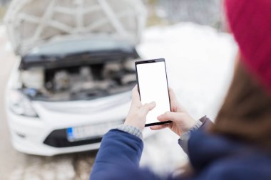 Kadının kırık arabaya karşı boş ekranlı akıllı telefonu tutmasını kapat. Kış sahnesi.