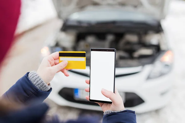 Close up of female holding blank screen smart phone and roadside assistance insurance membership card against broken car. Winter scene.