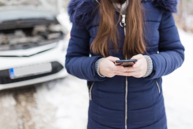 Kadın kış günü kaputu açık arabaya karşı akıllı telefon kullanıyor. Yol kenarı yardım hizmeti kavramı.