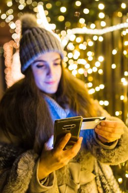 Dışarıda Noel ışıkları karşısında elinde cep telefonu ve kredi kartı tutan genç bir kadın. Tatil çevrimiçi alışveriş kavramı.