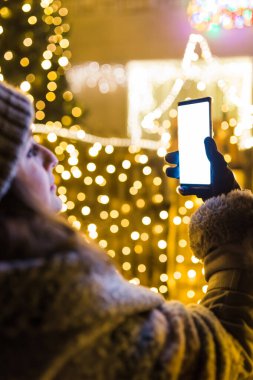 Yeni yıl dekorasyon lambalarıyla evin önünde boş ekran akıllı telefon kullanan bir kadın..