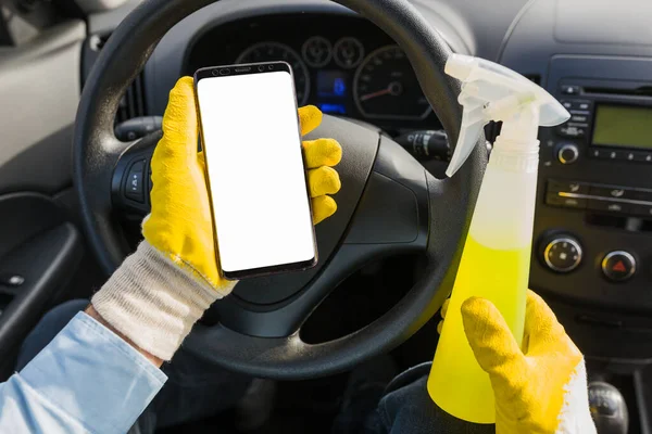 Close up of car wash worker holding blank screen smart phone and disinfectant spray bottle against car dashboard.