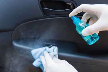 Erkek, koruyucu eldivenlerle arabanın içini temizlerken mikrofiber bez ve bir şişe sprey dezenfektan kullanıyor..