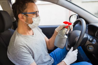 Şoför cerrahi maske ve koruyucu eldiven takarken enfeksiyonu önlemek için direksiyona dezenfektan sıkıyor..