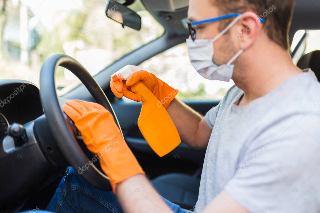 Worker with face mask cleaning car steering wheel with microfiber cloth and holding spray bottle of disinfectant.