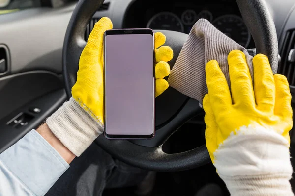 Close up of car wash worker holding blank screen smart phone and microfiber cloth against steering wheel
