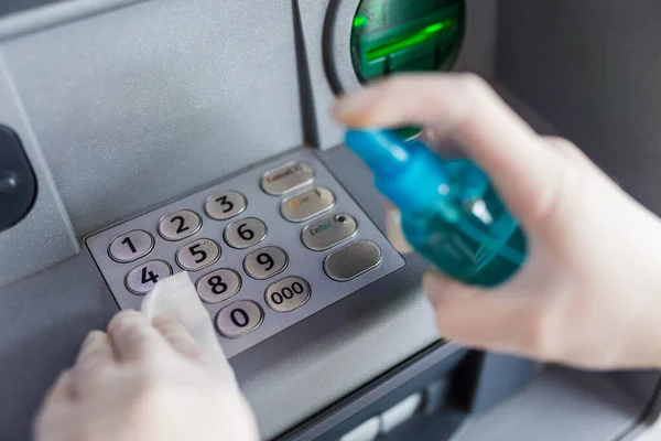 Hands in surgical rubber gloves sanitizing atm machine buttons with wet wipe and spray bottle disinfectant, covid-19 prevention.