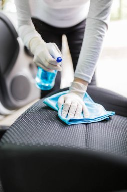 Koruyucu eldivenli bir kadın mikrofiber bez ve sprey dezenfektanla araba koltuğunu temizliyor..