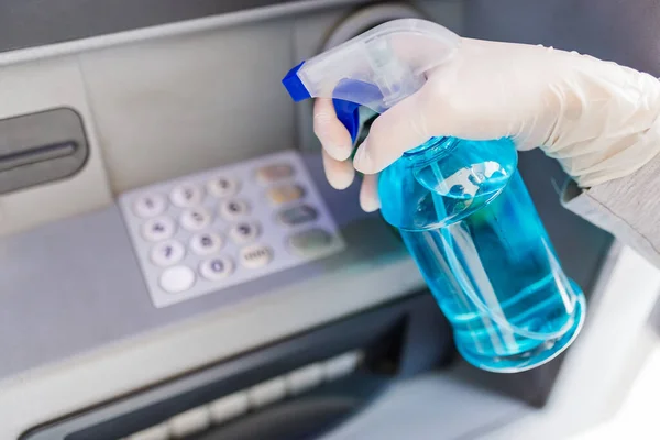 Close up of hand wearing surgical glove and holding bottle of cleaning alcohol to disinfect ATM numeric keyboard before withdrawing cash. Coronavirus concept.