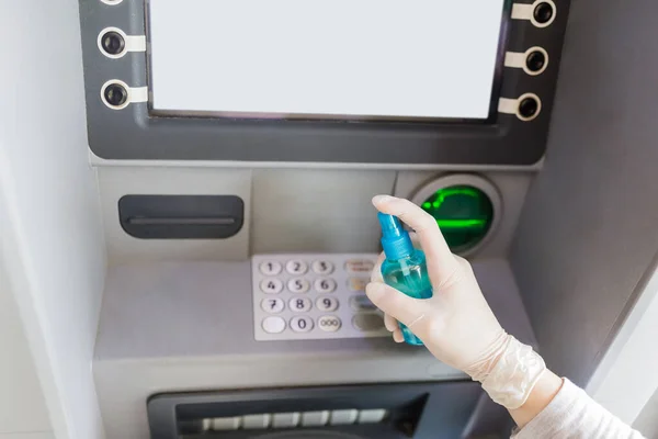 Close up of hand wearing medical glove and holding bottle of cleaning alcohol to disinfect ATM numeric keyboard before withdrawing cash. Coronavirus prevention concept.
