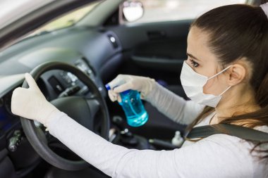 Yüz maskeli ve lastik eldivenli bir kadın araba direksiyonunu bakterilerden korumak için sprey şişe dezenfektanı kullanıyor. Antiseptik, hijyen ve sağlık konsepti.