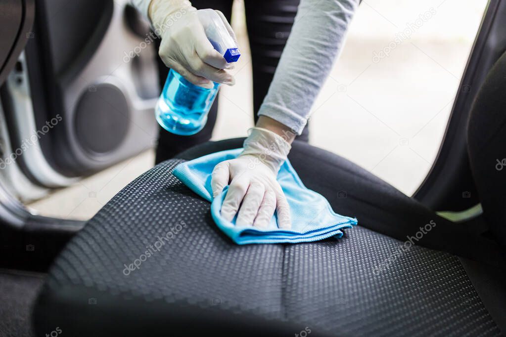 Close up of female wearing surgical gloves while cleaning car interior with disinfection liquid sprayer and microfiber cloth.