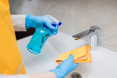 Ellerinde mikrofiber bez tutan lastik eldivenler ve lavabo musluğunu dezenfekte etmek için sprey şişesi dezenfekte eden kadın eller..