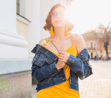 Çiçek tutan güzel bir kadının parlak portresi, sarı elbise, kot ceket, hippi tarzı, yaz modası, gülümseme, moda güneş gözlüğü.