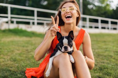 Turuncu elbiseli, gülümseyen genç kadın parkta köpekle oynarken eğleniyor, yaz stili, neşeli bir ruh hali.