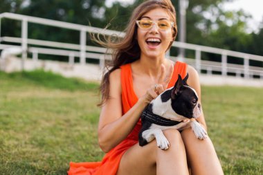 Turuncu elbiseli, gülümseyen genç kadın parkta köpekle oynarken eğleniyor, yaz stili, neşeli bir ruh hali.