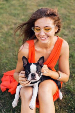 Yazlık parkta çimenlerde oturan, Boston Terrier köpeği tutan, gülümseyen, turuncu elbise giyen, moda tarzı olan, güneş gözlüklü, evcil hayvanla oynayan, eğlenen mutlu bir kadının portresi.
